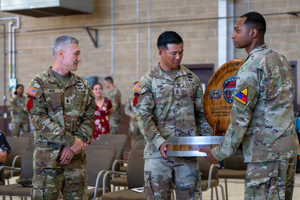 Three U.S. Army soldiers in OCP (Operational Camouflage Pattern) uniforms are participating in what appears to be a military ceremony or award presentation. A wooden plaque or commemorative item is being presented, bearing unit insignia and the text referencing '3-501 Assault Helicopter Battalion, Combat Aviation' and 'CSM STEVE' with a date of September 2023. The event is taking place indoors in what appears to be a gymnasium or community hall on a military installation.