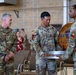 Three U.S. Army soldiers in OCP (Operational Camouflage Pattern) uniforms are participating in what appears to be a military ceremony or award presentation. A wooden plaque or commemorative item is being presented, bearing unit insignia and the text referencing '3-501 Assault Helicopter Battalion, Combat Aviation' and 'CSM STEVE' with a date of September 2023. The event is taking place indoors in what appears to be a gymnasium or community hall on a military installation.