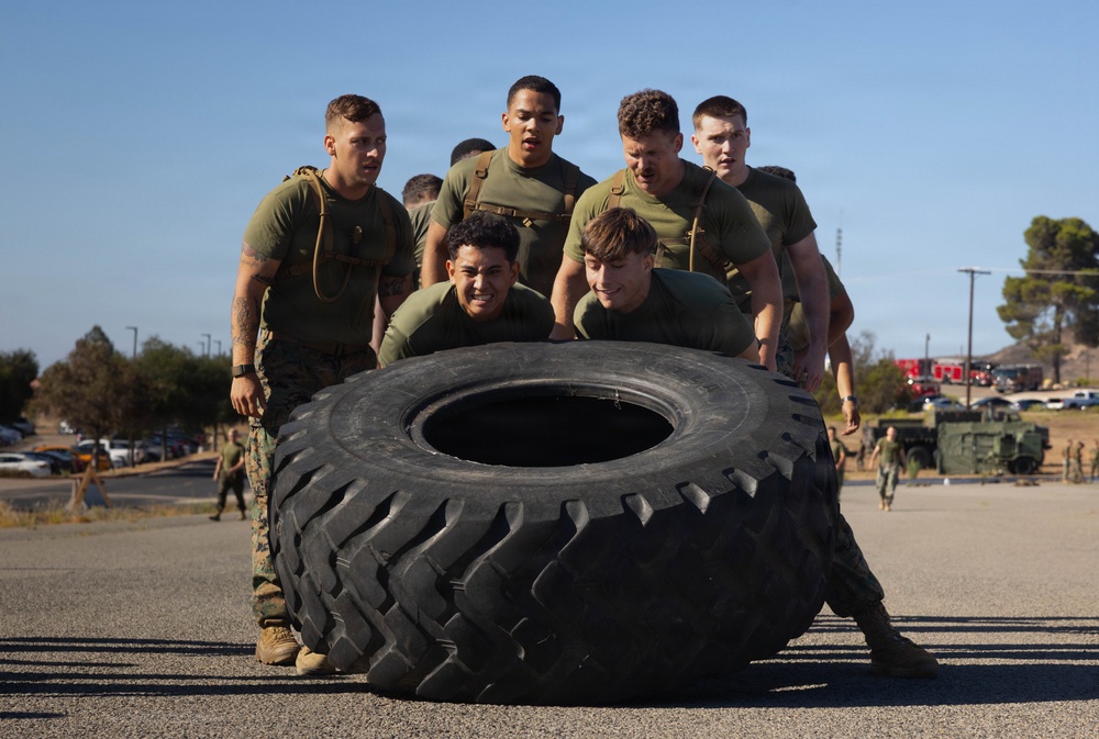 A group of military personnel in olive drab uniforms and tactical vests are collectively pushing a large heavy-equipment tire across a paved surface. This appears to be a physical training or team-building exercise conducted at a military installation. The activity requires coordinated group effort and is consistent with standard military fitness training drills.