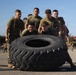 A group of military personnel in olive drab uniforms and tactical vests are collectively pushing a large heavy-equipment tire across a paved surface. This appears to be a physical training or team-building exercise conducted at a military installation. The activity requires coordinated group effort and is consistent with standard military fitness training drills.