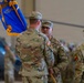 A U.S. Army military ceremony, likely a change of command or activation ceremony, taking place inside a hangar. Soldiers in OCP (Operational Camouflage Pattern) uniforms are participating in a guidon/flag transfer, with one soldier holding a blue unit flag with gold fringe. The setting includes a helicopter visible in the background and additional personnel observing the ceremony.