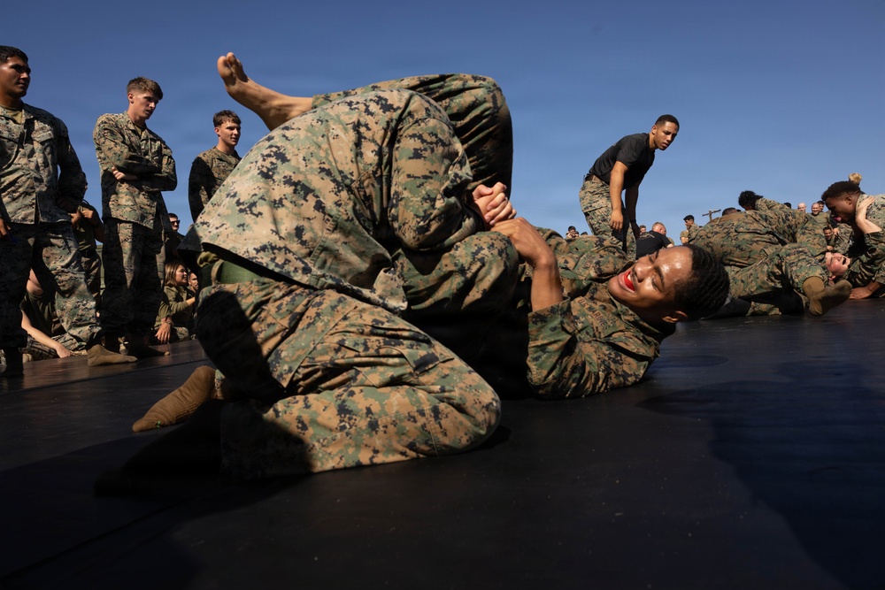 Military personnel in MARPAT (Marine pattern) camouflage uniforms are engaged in ground combat/grappling training on a black mat surface outdoors. Two individuals in the foreground are actively sparring in what appears to be a submission hold or joint lock technique. Multiple other pairs of personnel are training simultaneously in the background, observed by standing onlookers.