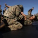 Military personnel in MARPAT (Marine pattern) camouflage uniforms are engaged in ground combat/grappling training on a black mat surface outdoors. Two individuals in the foreground are actively sparring in what appears to be a submission hold or joint lock technique. Multiple other pairs of personnel are training simultaneously in the background, observed by standing onlookers.