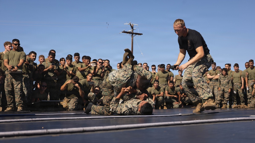 A group of military personnel, likely U.S. Marines based on their camouflage uniforms, are gathered around a makeshift ring or mat watching a combat/martial arts demonstration. Two individuals in camouflage uniforms are grappling on the ground while a third person in a black t-shirt stands over them in a dominant position. A large crowd of uniformed spectators surrounds the demonstration area, many recording with phones.