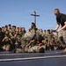A group of military personnel, likely U.S. Marines based on their camouflage uniforms, are gathered around a makeshift ring or mat watching a combat/martial arts demonstration. Two individuals in camouflage uniforms are grappling on the ground while a third person in a black t-shirt stands over them in a dominant position. A large crowd of uniformed spectators surrounds the demonstration area, many recording with phones.