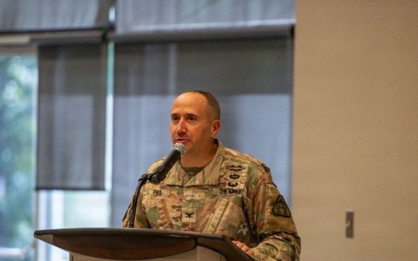 A U.S. Army officer in OCP (Operational Camouflage Pattern) uniform is delivering a speech at a podium bearing the Fort Belvoir 'Leaders in Excellence' seal. The setting appears to be an indoor conference or event hall with large windows in the background. The officer's rank insignia and name tape are partially visible on the uniform.