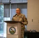A U.S. Army officer in OCP (Operational Camouflage Pattern) uniform is delivering a speech at a podium bearing the Fort Belvoir 'Leaders in Excellence' seal. The setting appears to be an indoor conference or event hall with large windows in the background. The officer's rank insignia and name tape are partially visible on the uniform.