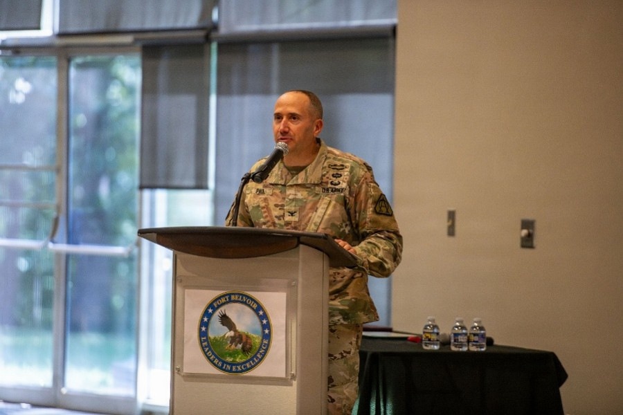 A U.S. Army officer in OCP (Operational Camouflage Pattern) uniform is delivering a speech at a podium bearing the Fort Belvoir 'Leaders in Excellence' seal. The setting appears to be an indoor conference or event hall with large windows in the background. The officer's rank insignia and name tape are partially visible on the uniform.