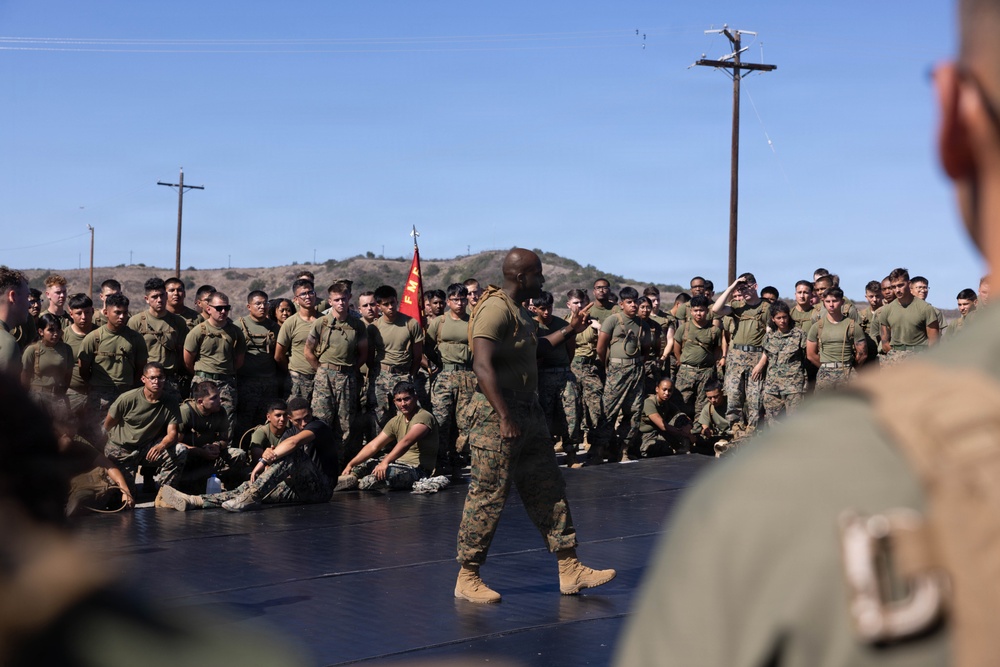 A military instructor or senior non-commissioned officer addresses a large formation of Marines on an outdoor training platform. The speaker stands centrally on a dark mat surface, gesturing with one hand while facing the assembled troops. The setting appears to be a military base, likely Camp Pendleton or a similar installation, based on the terrain and infrastructure visible.