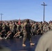 A military instructor or senior non-commissioned officer addresses a large formation of Marines on an outdoor training platform. The speaker stands centrally on a dark mat surface, gesturing with one hand while facing the assembled troops. The setting appears to be a military base, likely Camp Pendleton or a similar installation, based on the terrain and infrastructure visible.