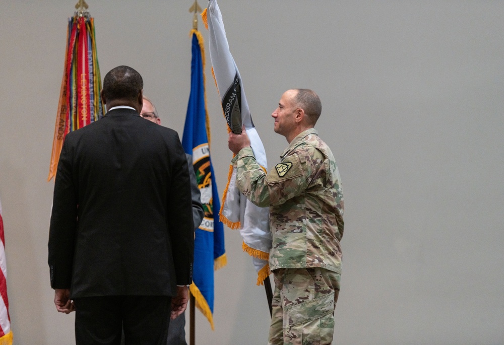 A military ceremony is taking place indoors, likely a change of command or unit activation event. A soldier in Army Combat Uniform (ACU/OCP camouflage) is unfurling or presenting a guidon/unit flag, while two civilians in dark suits stand nearby facing away from the camera. Multiple flags and streamers are visible in the background.