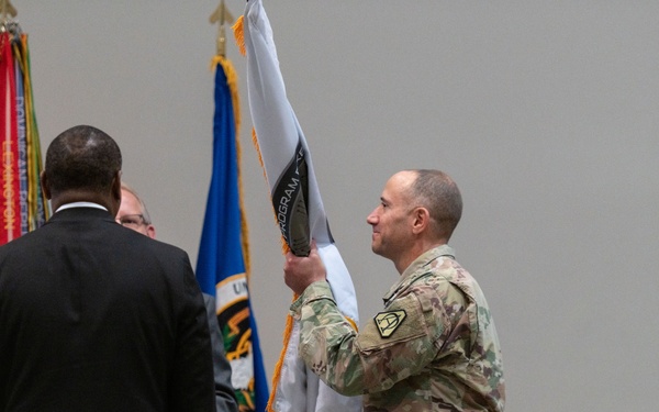 A military ceremony is taking place indoors, likely a change of command or unit activation event. A soldier in Army Combat Uniform (ACU/OCP camouflage) is unfurling or presenting a guidon/unit flag, while two civilians in dark suits stand nearby facing away from the camera. Multiple flags and streamers are visible in the background.