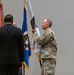 A military ceremony is taking place indoors, likely a change of command or unit activation event. A soldier in Army Combat Uniform (ACU/OCP camouflage) is unfurling or presenting a guidon/unit flag, while two civilians in dark suits stand nearby facing away from the camera. Multiple flags and streamers are visible in the background.