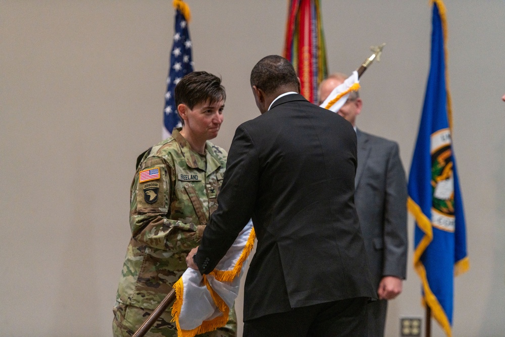 A military ceremony is taking place indoors, likely a change of command or activation/deactivation ceremony. A female soldier wearing U.S. Army combat uniform with the 101st Airborne Division patch and name tape 'FREELAND' is exchanging or receiving a furled guidon/flag with a civilian in a dark suit. Several flags, including the U.S. flag and what appears to be a Department of the Army or state flag, are visible in the background.