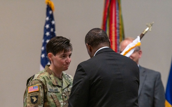 A military ceremony is taking place indoors, likely a change of command or activation/deactivation ceremony. A female soldier wearing U.S. Army combat uniform with the 101st Airborne Division patch and name tape 'FREELAND' is exchanging or receiving a furled guidon/flag with a civilian in a dark suit. Several flags, including the U.S. flag and what appears to be a Department of the Army or state flag, are visible in the background.