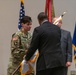 A military ceremony is taking place indoors, likely a change of command or activation/deactivation ceremony. A female soldier wearing U.S. Army combat uniform with the 101st Airborne Division patch and name tape 'FREELAND' is exchanging or receiving a furled guidon/flag with a civilian in a dark suit. Several flags, including the U.S. flag and what appears to be a Department of the Army or state flag, are visible in the background.