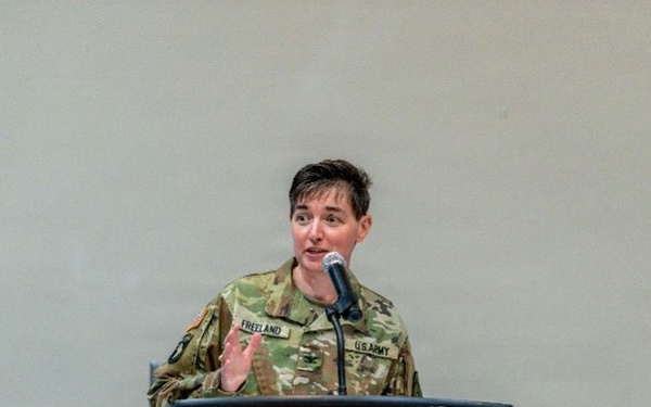 A U.S. Army soldier named Freeland is speaking at a podium during an event at Fort Belvoir. The individual is wearing Army Combat Uniform (ACU) in OCP (Operational Camouflage Pattern) and is gesturing while addressing an audience. The Fort Belvoir seal is visible on the front of the podium.