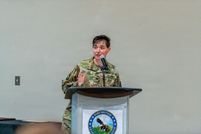 A U.S. Army soldier named Freeland is speaking at a podium during an event at Fort Belvoir. The individual is wearing Army Combat Uniform (ACU) in OCP (Operational Camouflage Pattern) and is gesturing while addressing an audience. The Fort Belvoir seal is visible on the front of the podium.