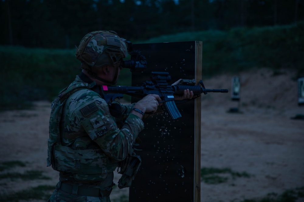 A U.S. Army soldier in full combat gear is conducting a nighttime live-fire training exercise at a shooting range. The soldier is using a barrier for cover while aiming a rifle, with visible targets downrange. The low-light conditions suggest this is a night qualification or tactical shooting drill.