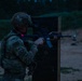 A U.S. Army soldier in full combat gear is conducting a nighttime live-fire training exercise at a shooting range. The soldier is using a barrier for cover while aiming a rifle, with visible targets downrange. The low-light conditions suggest this is a night qualification or tactical shooting drill.