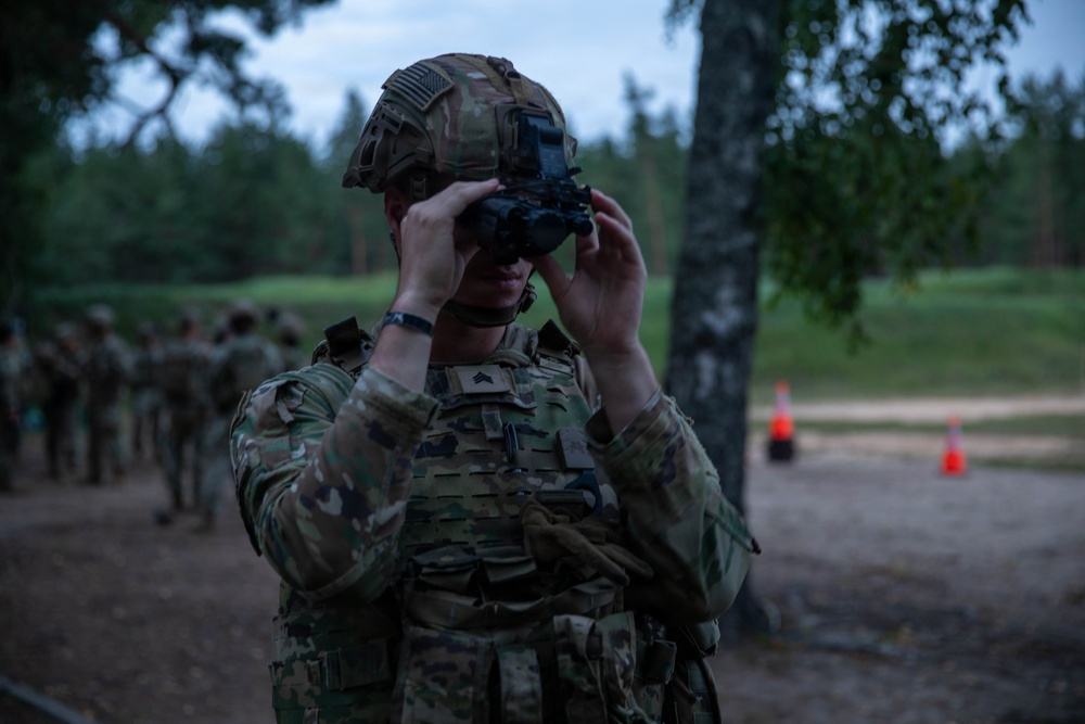 A U.S. Army soldier in full combat gear is adjusting or testing night vision goggles (NVGs) during what appears to be a dusk or low-light training exercise. The soldier wears Operational Camouflage Pattern (OCP) uniform, a tactical helmet with a U.S. flag patch, and a plate carrier vest. Additional soldiers in formation are visible in the background, suggesting a unit-level training event.