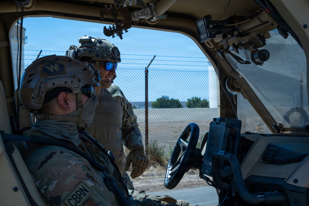 Two military personnel in full tactical gear are interacting near a military vehicle, with one seated inside and one standing at the open door. Both wear combat helmets with accessories and MultiCam/OCP pattern uniforms. The setting appears to be a secured perimeter area with chain-link fencing topped with barbed wire visible in the background.