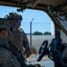 Two military personnel in full tactical gear are interacting near a military vehicle, with one seated inside and one standing at the open door. Both wear combat helmets with accessories and MultiCam/OCP pattern uniforms. The setting appears to be a secured perimeter area with chain-link fencing topped with barbed wire visible in the background.