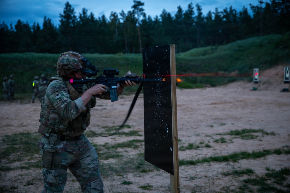 A soldier in MultiCam/OCP camouflage uniform is conducting a live-fire night shooting exercise at a military range during dusk or low-light conditions. The soldier is firing an M4/M16-style rifle equipped with a night vision device and laser aiming module, with a visible red laser beam striking a black target board. Additional soldiers and range targets are visible in the background.