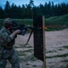 A soldier in MultiCam/OCP camouflage uniform is conducting a live-fire night shooting exercise at a military range during dusk or low-light conditions. The soldier is firing an M4/M16-style rifle equipped with a night vision device and laser aiming module, with a visible red laser beam striking a black target board. Additional soldiers and range targets are visible in the background.