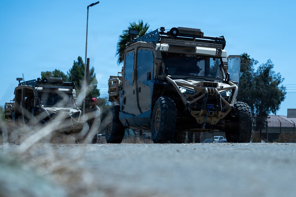 The image shows two tactical military utility vehicles in a tan/desert camouflage color scheme parked or moving on a dusty surface. The vehicles appear to be heavily modified off-road platforms with armored cab enclosures, roof-mounted equipment racks, and light bars. The setting appears to be a military installation or training area in an arid environment.