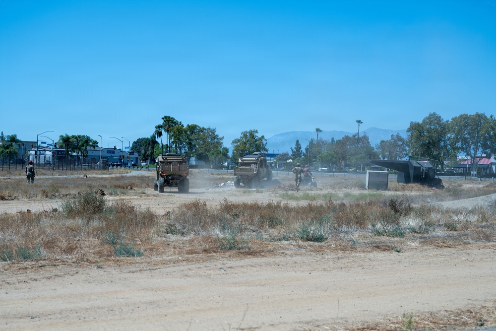 The image shows a dry, dusty open field with multiple military-style vehicles operating in a convoy or work formation. Personnel in military attire are visible near the vehicles, which are kicking up dust as they move across the arid terrain. The scene appears to take place near an urban area, with trees, street lights, and buildings visible in the background.