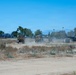 The image shows a dry, dusty open field with multiple military-style vehicles operating in a convoy or work formation. Personnel in military attire are visible near the vehicles, which are kicking up dust as they move across the arid terrain. The scene appears to take place near an urban area, with trees, street lights, and buildings visible in the background.