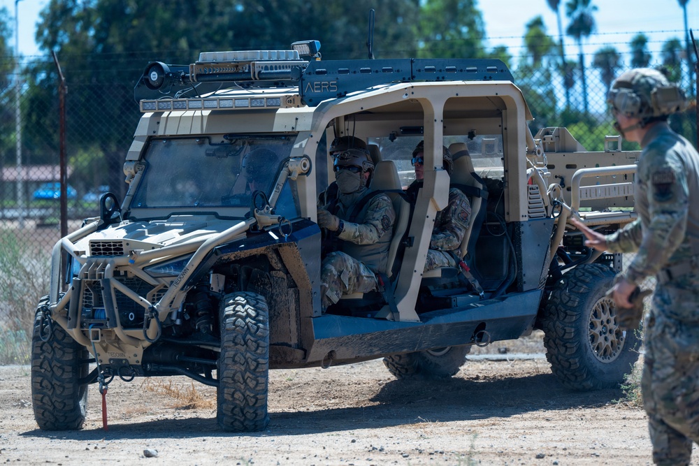 The image shows military personnel operating a tactical utility vehicle (UTV) in what appears to be a training or operational environment. The vehicle, marked with 'AERS' on its roof rack, is a tan/desert-colored off-road platform equipped with roof-mounted sensors, communications equipment, and a winch. Two soldiers are seated inside the vehicle while a third stands nearby, appearing to provide direction or instruction.