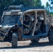 The image shows military personnel operating a tactical utility vehicle (UTV) in what appears to be a training or operational environment. The vehicle, marked with 'AERS' on its roof rack, is a tan/desert-colored off-road platform equipped with roof-mounted sensors, communications equipment, and a winch. Two soldiers are seated inside the vehicle while a third stands nearby, appearing to provide direction or instruction.