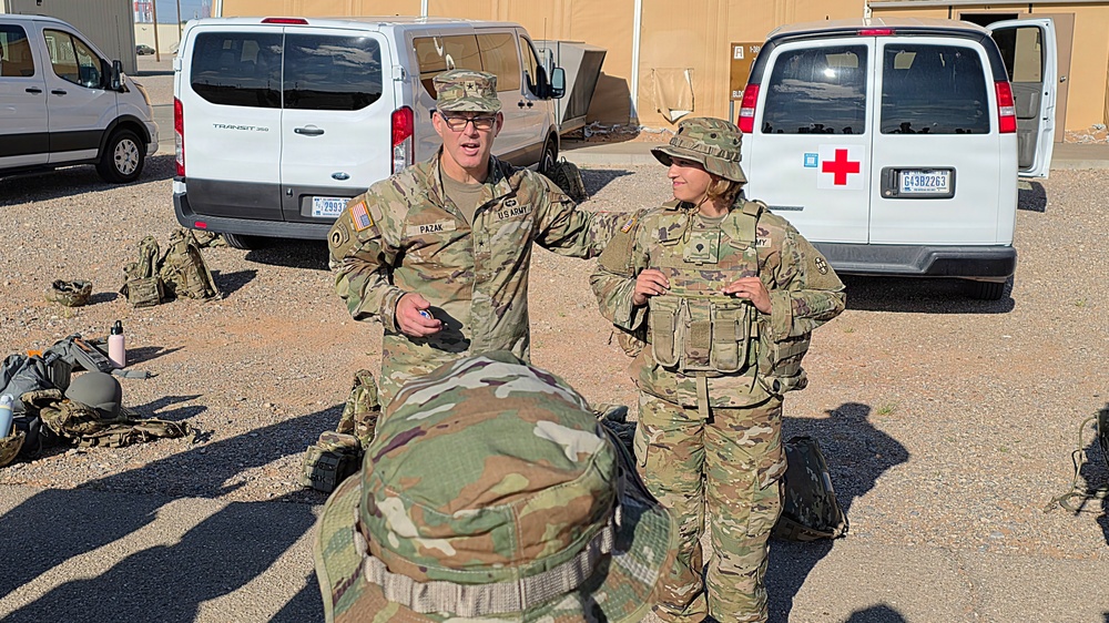 Two U.S. Army soldiers in OCP (Operational Camouflage Pattern) uniforms are conversing in a gravel lot at what appears to be a military installation. The taller soldier is identified by a name tape reading 'PAZAK' and both wear U.S. Army insignia. A medical (Red Cross) vehicle and a Ford Transit 350 van are parked in the background.