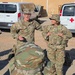 Two U.S. Army soldiers in OCP (Operational Camouflage Pattern) uniforms are conversing in a gravel lot at what appears to be a military installation. The taller soldier is identified by a name tape reading 'PAZAK' and both wear U.S. Army insignia. A medical (Red Cross) vehicle and a Ford Transit 350 van are parked in the background.