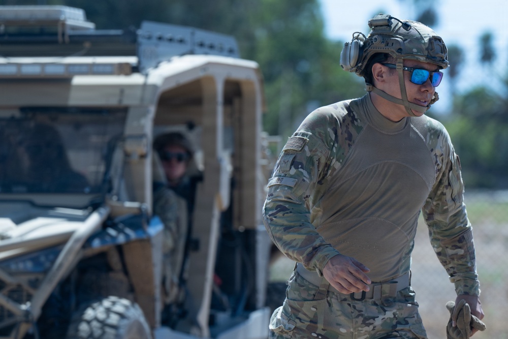 Two military personnel are visible in an outdoor field environment. The foreground subject is wearing full tactical gear including a combat helmet with communications equipment, OCP/MultiCam camouflage uniform, blue mirrored sunglasses, and a tactical belt. A second individual is seated in a tan/desert-colored utility tactical vehicle (UTV) in the background.