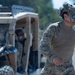Two military personnel are visible in an outdoor field environment. The foreground subject is wearing full tactical gear including a combat helmet with communications equipment, OCP/MultiCam camouflage uniform, blue mirrored sunglasses, and a tactical belt. A second individual is seated in a tan/desert-colored utility tactical vehicle (UTV) in the background.