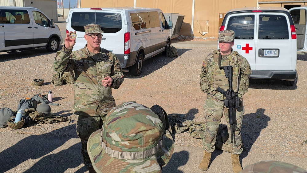 A U.S. Army officer, identified by nametape as 'PAZAK', is holding up what appears to be a challenge coin while addressing personnel in a military compound. A second soldier stands at ease holding a rifle to the right. The setting appears to be a forward operating base or military installation, with vehicles including a medical (Red Cross) van visible in the background.