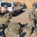 A U.S. Army officer, identified by nametape as 'PAZAK', is holding up what appears to be a challenge coin while addressing personnel in a military compound. A second soldier stands at ease holding a rifle to the right. The setting appears to be a forward operating base or military installation, with vehicles including a medical (Red Cross) van visible in the background.