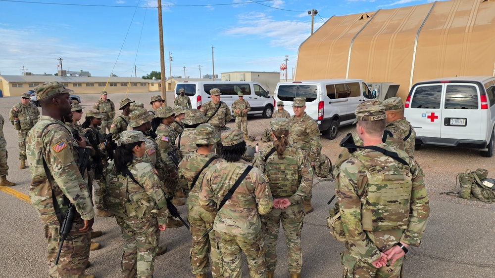 A group of U.S. Army soldiers in OCP (Operational Camouflage Pattern) uniforms are gathered in a formation huddle on a military installation, appearing to receive a briefing or instructions from a leader. The setting includes military vehicles, including a Red Cross-marked van, a large tan tent structure, and standard military base buildings in the background. The scene suggests a pre-mission briefing or training exercise.