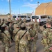 A group of U.S. Army soldiers in OCP (Operational Camouflage Pattern) uniforms are gathered in a formation huddle on a military installation, appearing to receive a briefing or instructions from a leader. The setting includes military vehicles, including a Red Cross-marked van, a large tan tent structure, and standard military base buildings in the background. The scene suggests a pre-mission briefing or training exercise.
