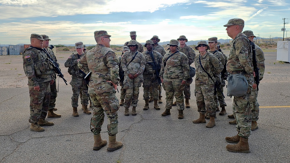 A group of approximately 15-20 U.S. Army soldiers in MultiCam/OCP camouflage uniforms are gathered in a formation or briefing circle on what appears to be a paved outdoor area, likely a military installation. A leader or senior soldier appears to be addressing the group. The setting appears to be an arid or desert environment based on the terrain and vegetation visible in the background.