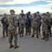 A group of approximately 15-20 U.S. Army soldiers in MultiCam/OCP camouflage uniforms are gathered in a formation or briefing circle on what appears to be a paved outdoor area, likely a military installation. A leader or senior soldier appears to be addressing the group. The setting appears to be an arid or desert environment based on the terrain and vegetation visible in the background.