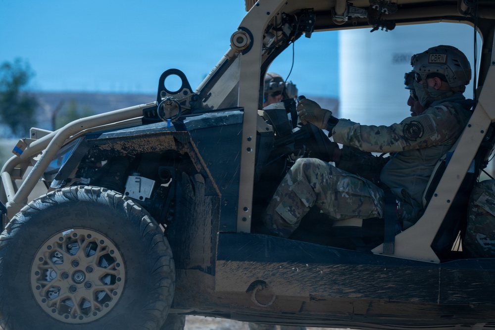 Two military personnel in full combat gear are operating a tactical utility vehicle (UTV), likely a Polaris MRZR or similar military off-road vehicle. The driver wears a helmet marked 'CBRN' (Chemical, Biological, Radiological, and Nuclear), indicating a specialized military role. The vehicle shows signs of heavy use with mud and dirt visible on its exterior.