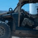 Two military personnel in full combat gear are operating a tactical utility vehicle (UTV), likely a Polaris MRZR or similar military off-road vehicle. The driver wears a helmet marked 'CBRN' (Chemical, Biological, Radiological, and Nuclear), indicating a specialized military role. The vehicle shows signs of heavy use with mud and dirt visible on its exterior.