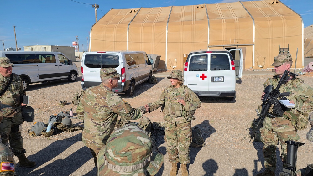 A group of U.S. Army soldiers in OCP (Operational Camouflage Pattern) uniforms are gathered in an outdoor area at what appears to be a military installation or training base. Two soldiers are visibly shaking hands in the center of the image, suggesting a greeting, recognition, or after-action interaction. The setting includes military support vehicles, including a white medical van marked with a Red Cross, and a large tan fabric structure in the background.