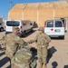 A group of U.S. Army soldiers in OCP (Operational Camouflage Pattern) uniforms are gathered in an outdoor area at what appears to be a military installation or training base. Two soldiers are visibly shaking hands in the center of the image, suggesting a greeting, recognition, or after-action interaction. The setting includes military support vehicles, including a white medical van marked with a Red Cross, and a large tan fabric structure in the background.