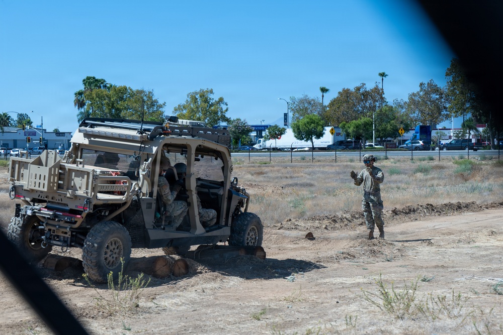 Military personnel are operating a tan/desert-colored tactical utility vehicle (UTV) on an unpaved dirt area near what appears to be a suburban roadway. One soldier is standing outside the vehicle gesturing, possibly directing the driver, while another remains seated inside. The setting appears to be a dry, open lot adjacent to a busy commercial street in a warm-climate urban area.
