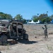 Military personnel are operating a tan/desert-colored tactical utility vehicle (UTV) on an unpaved dirt area near what appears to be a suburban roadway. One soldier is standing outside the vehicle gesturing, possibly directing the driver, while another remains seated inside. The setting appears to be a dry, open lot adjacent to a busy commercial street in a warm-climate urban area.